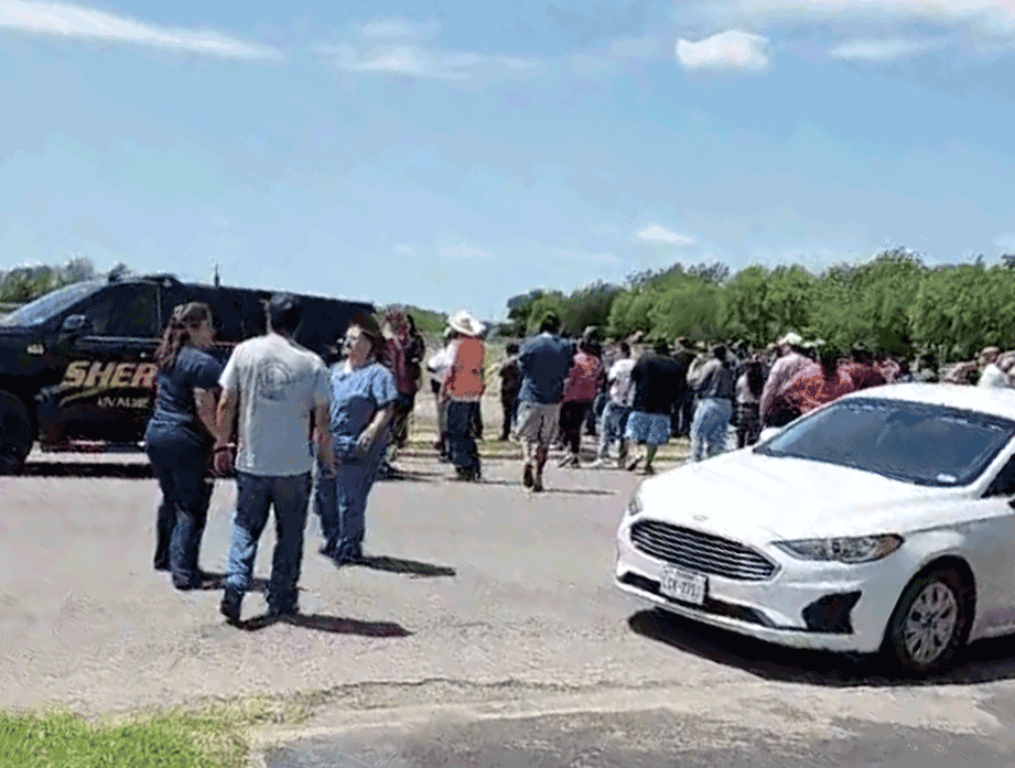 A photo of a crowd of people standing on a street outside the school. A black car with the word “Sheriff” on the side is also parked on the street.