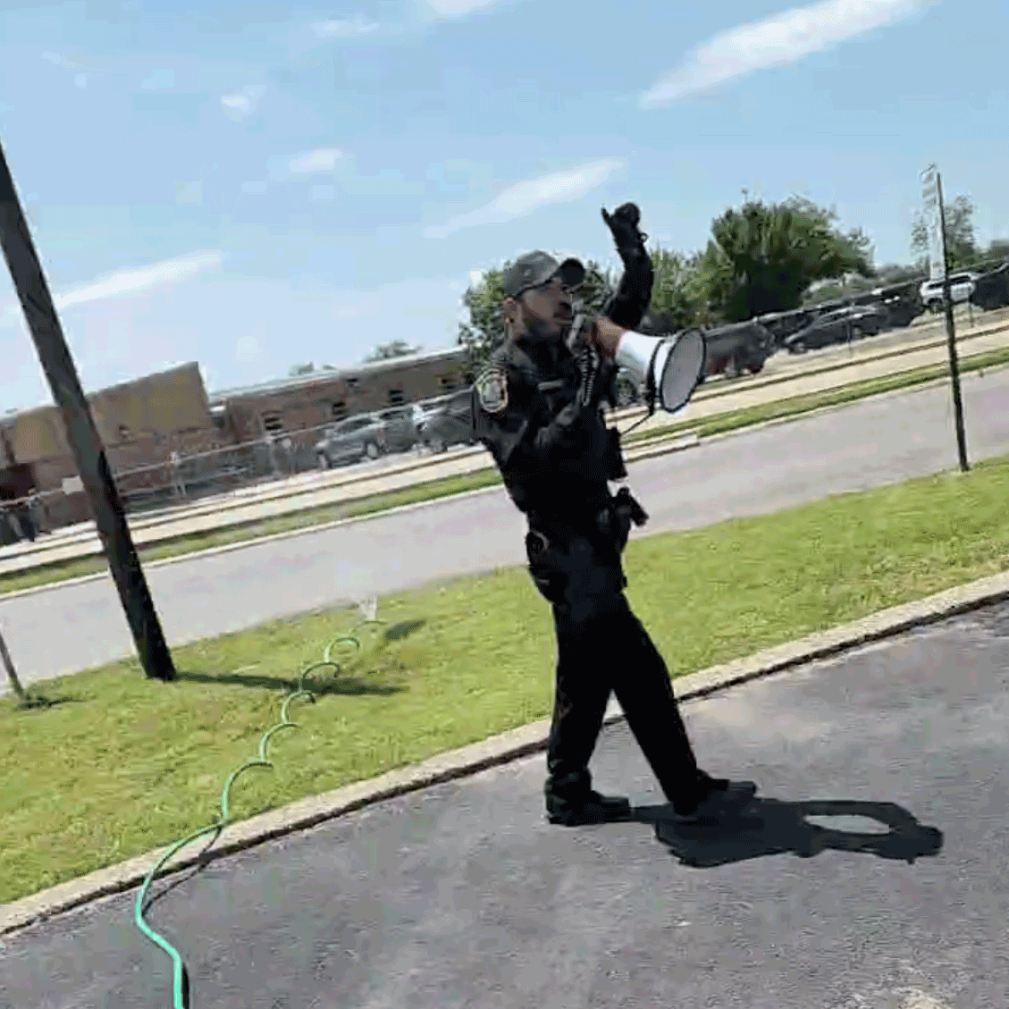 A photo of a police officer in uniform walking along a street outside of the school. He is speaking into a megaphone and gesturing toward the sky.