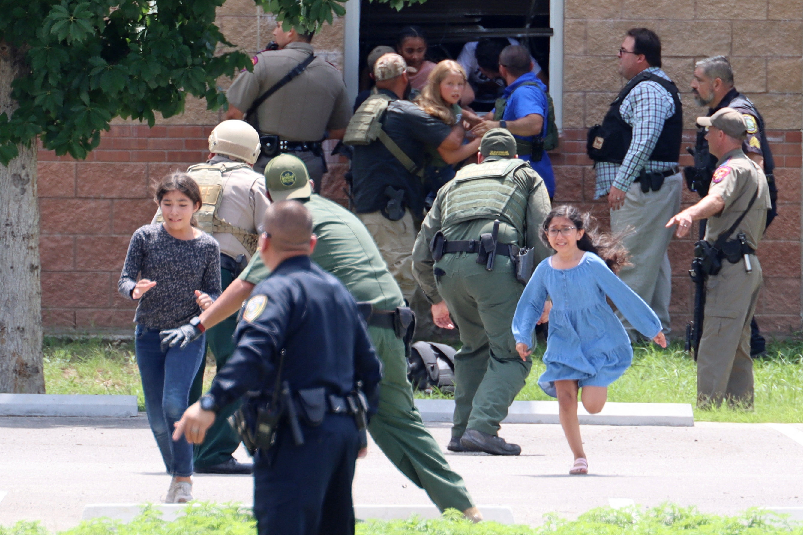 A photo showing the outside of a school building. Several children are being helped out of a first floor window by two to three adults. A group of other adults, several in uniform, are standing nearby. Two children are running from the direction of the school window toward the photographer.