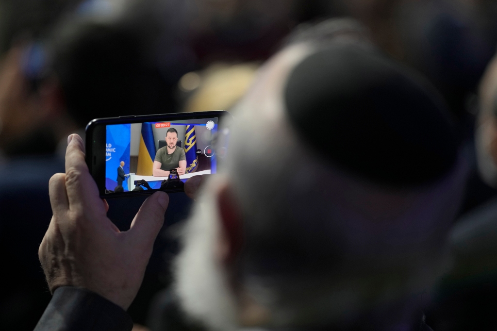 A man watches on his phone Ukrainian President Volodymyr Zelenskyy addressing the audience from Kyiv on a screen during the World Economic Forum in Davos, Switzerland, Monday, May 23, 2022. 