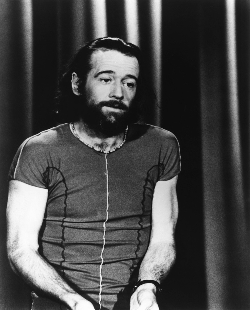 George Carlin performing on stage in the 1970s. He's got long hair and a beard and is wearing a short-sleeved shirt.
