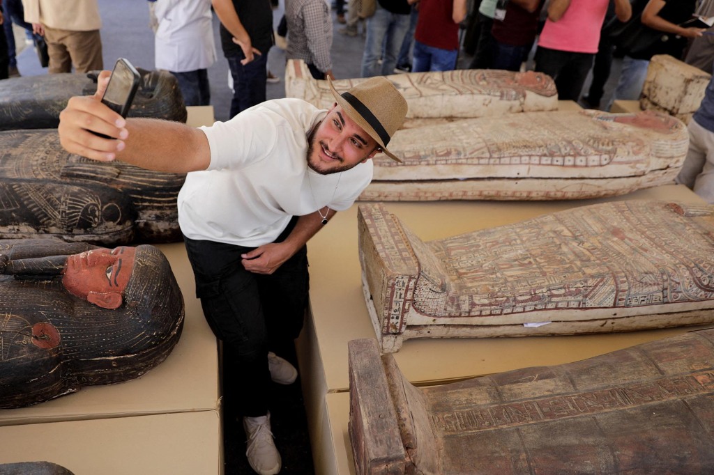 A man takes a selfie next to displayed sarcophagi.