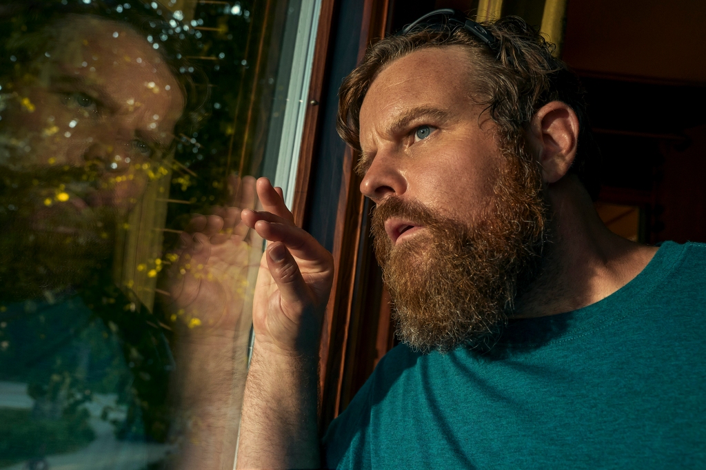Photo of Adam Bartley as Franklin and Irene's the nosy neighbor, Byron, who suspects something amiss. He's peering out of a window in his house. He has a bushy beard and is wearing a green T-shirt.