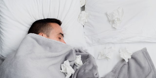 Ill young man in bed, covered with a light gray blanket, surrounded by used tissues