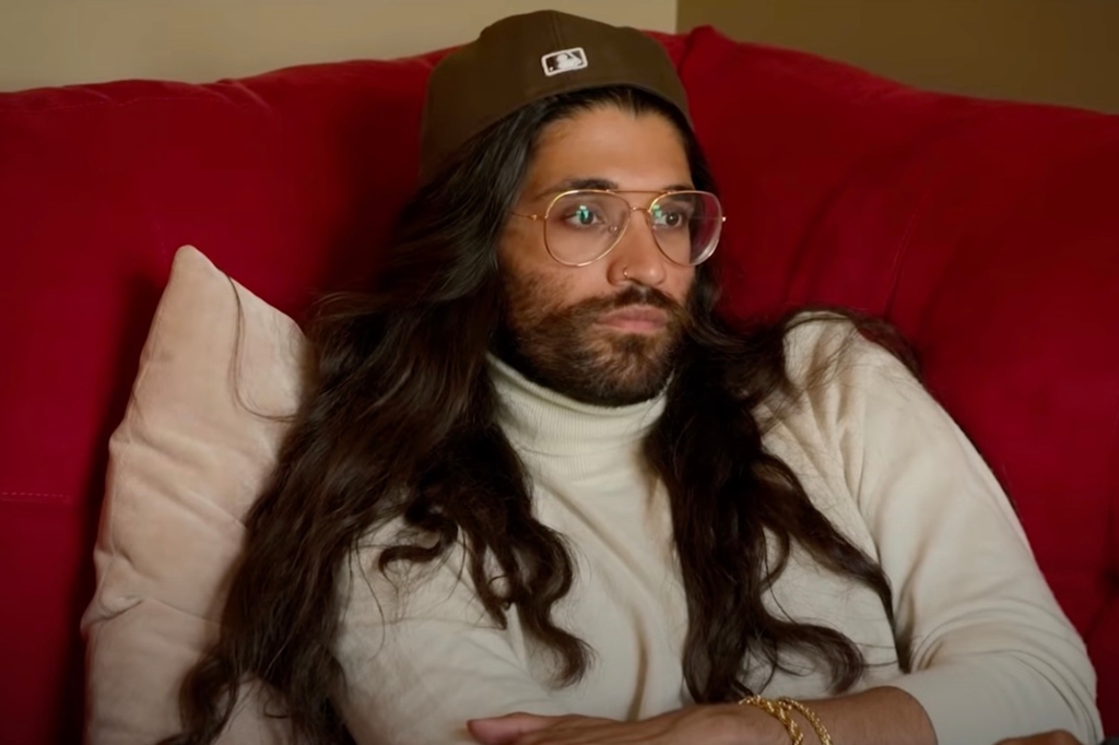 A long haired man lounges on a couch looking sad. 