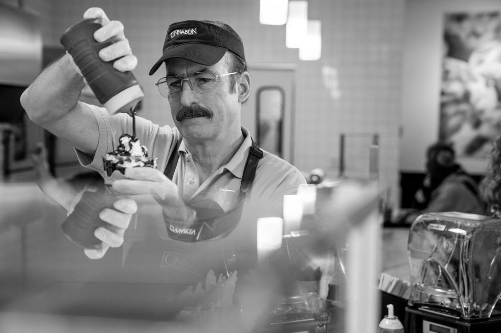 Bob Odenkirk as Saul Goodman/Gene Takavic. He's in the Cinnabon store and is pouring chocolate sauce over an ice cream sundae. He's wearing a hat with the word Cinnabon across the top. He's got a mustache and glasses.