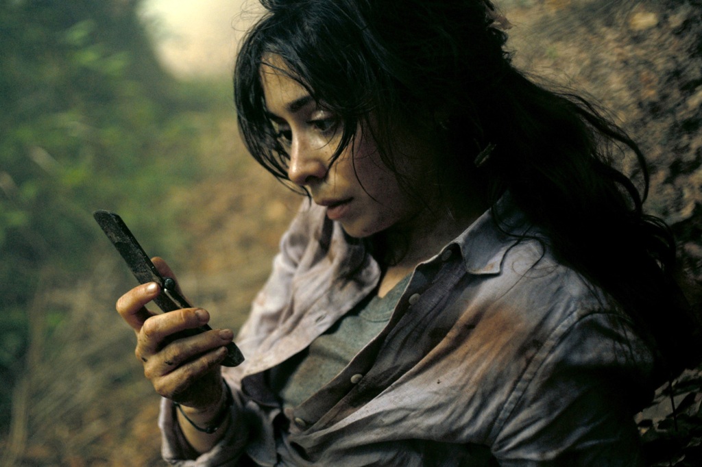 Emma (Cristin Milioti) sits in the woods looking at a phone. 