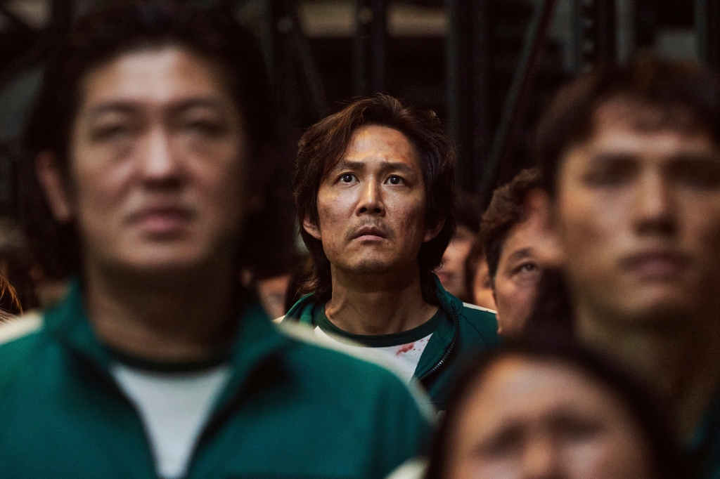 Photo of "Squid Game" star Lee Jung-jae in a scene from the Netflix series. He's in a crowd of people and looks frightened; there's a dab of blood on his shirt, which he's wearing underneath a green zip-up sweatshirt.