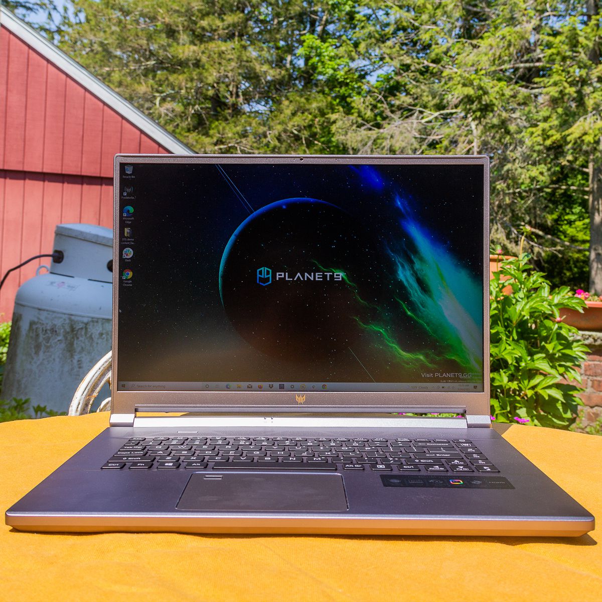 The Acer Predator Triton 500 SE on a yellow tablecloth in a garden, open. The screen displays the Planet9 logo on a dark background.