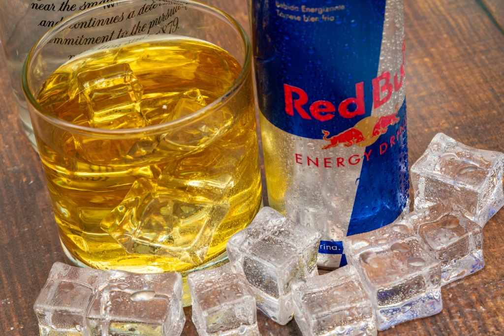 A redbull can sits on a bar next to a mixed drink.