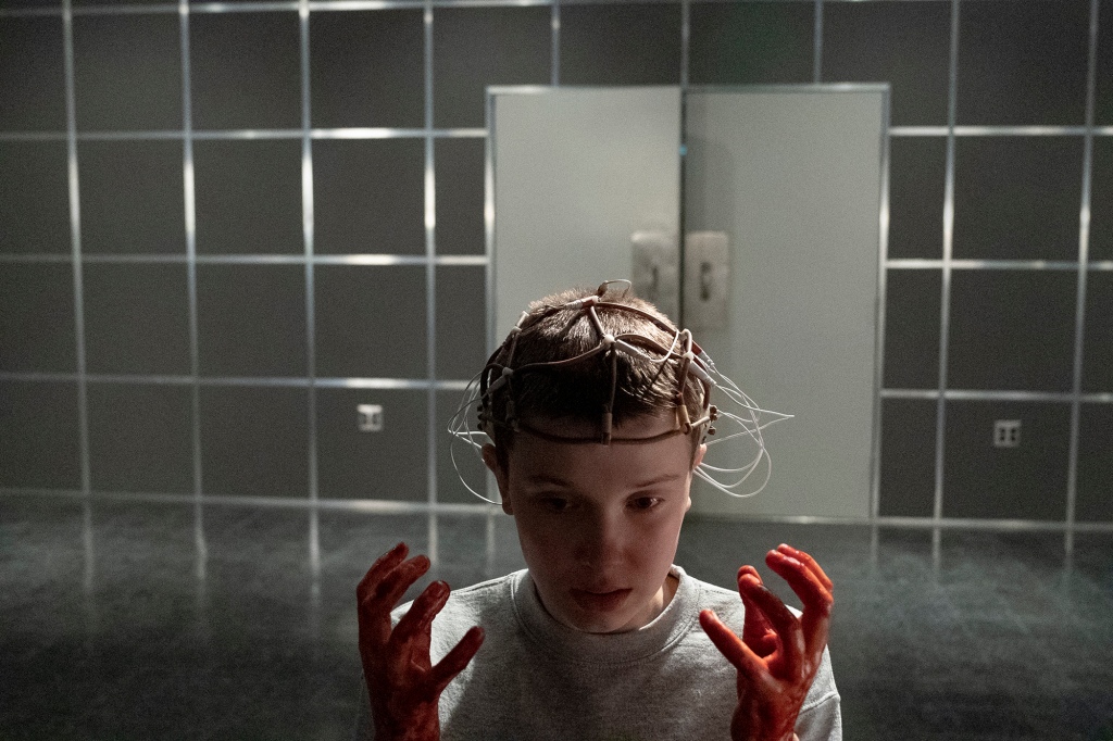 Eleven (Millie Bobby Brown) in a lab with a headpiece on and bloody hands. 
