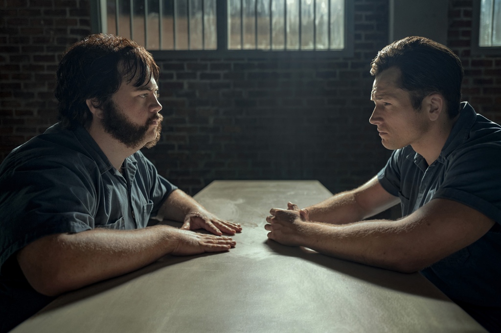 Photo of Paul Walter Hauser and Taron Egerton as Larry Hall and Jimmy Keene. They're seated at a table in prison facing each other and looking serious. They both have their hands on the table.