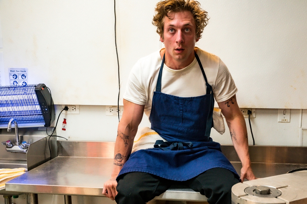Jeremy Allen White as as Carmen "Carmy" Berzatto. He's sitting on a table in the sandwich shop and is wearing a blue apron. He has a dumbfounded look on his face.