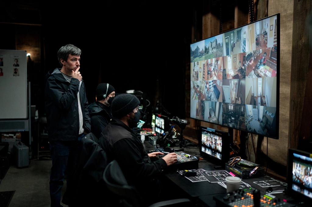 Nathan Fielder stands in front of a large screen and a set of computers. 