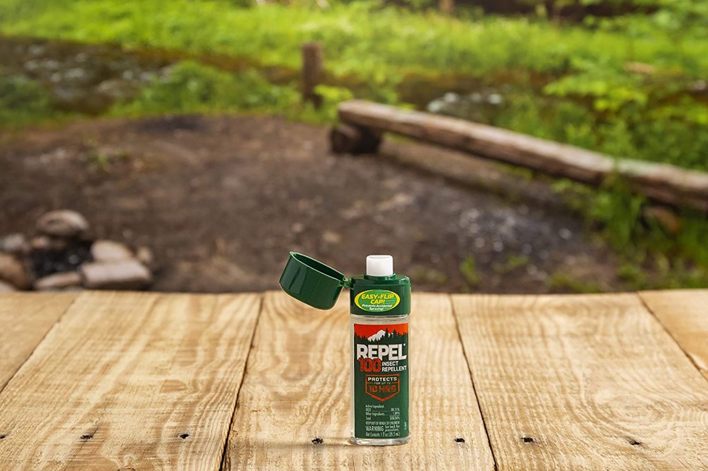 A mini container of bug spray sits on a wood floor with a background of a forrest 
