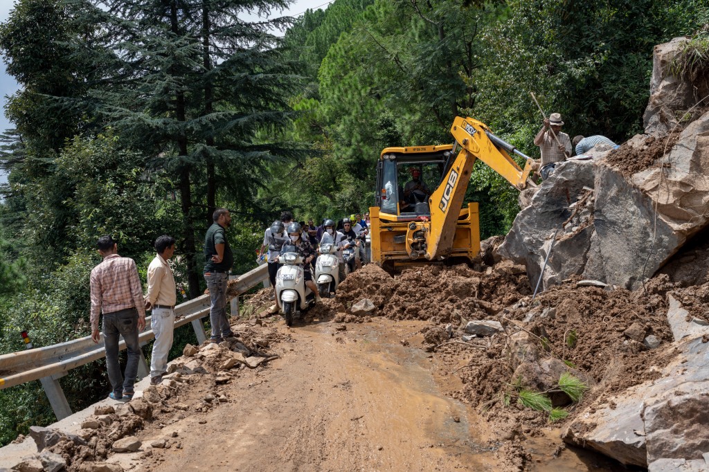 Scooter riders rush past an earthmover clearing a road of a big rock that came down with mud and plant debris in Dharmsala, India on Aug. 21, 2022. 