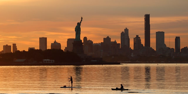 Two people paddle past the Statue of Liberty and the skyline of Brooklyn as the sun rises in New York City on August 15, 2022, as seen from Jersey City. 