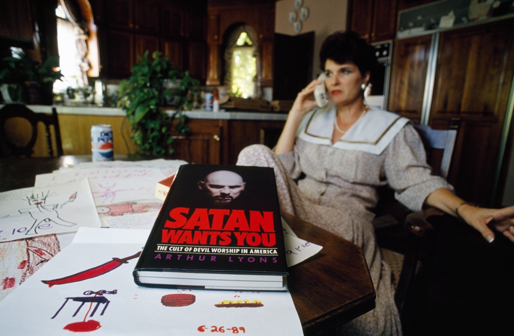 Photo of Faye Yager in the 1980s. She's talking on the phone and there's a book on the table in front of her entitled "Satan Wants You: The Cult of Devil Worship in America."