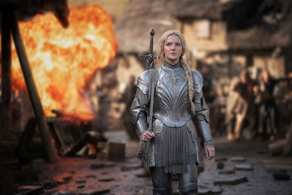 Galadriel (Morfydd Clark) wearing armor holding a sword with fire in the background. 