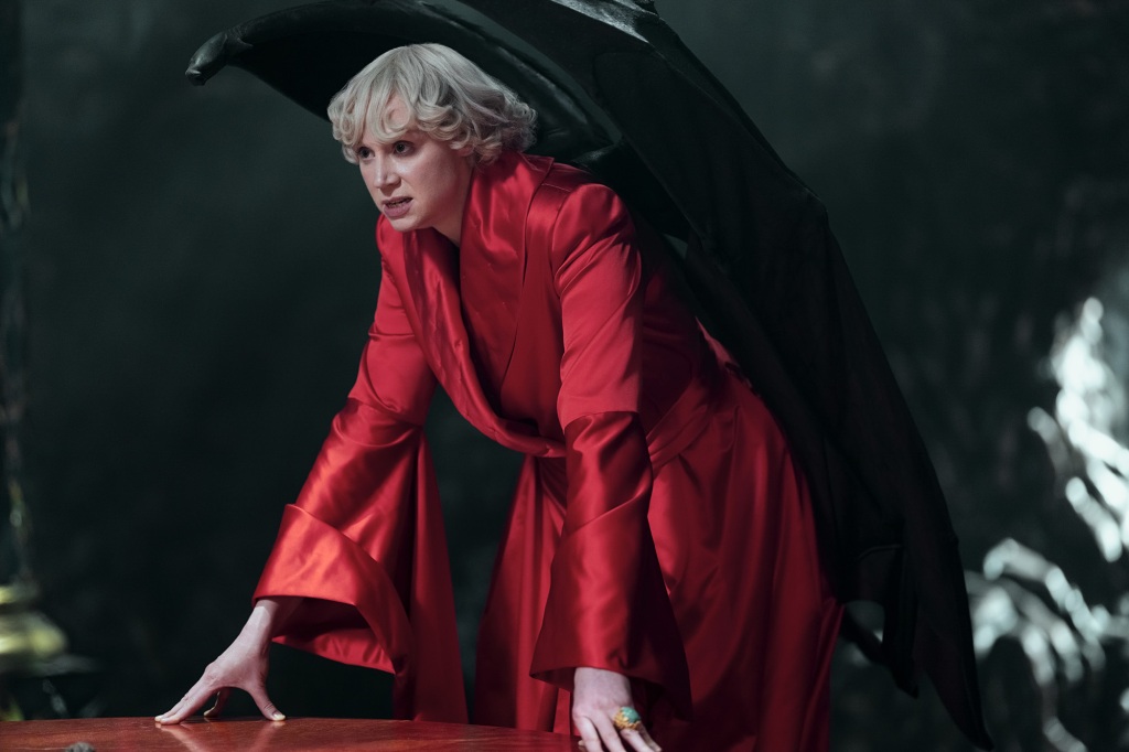 Gwendoline Christie as Lucifer in "The Sandman" leaning over a table in a  red robe. 