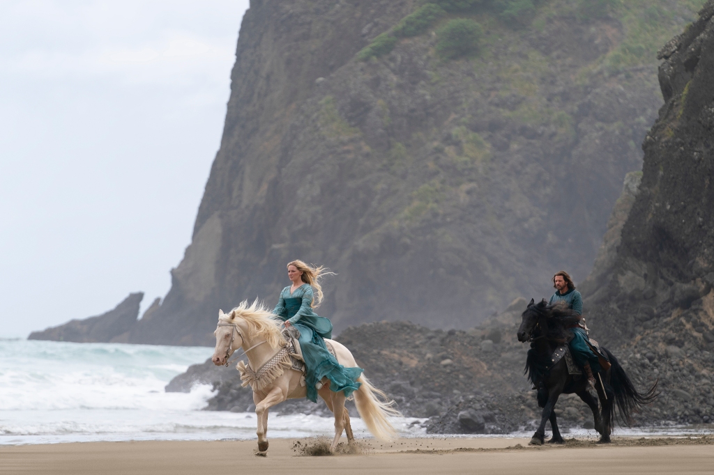 Lloyd Owen on a horse with Morfydd Clark also on a horse, riding on a beach by cliffs. 
