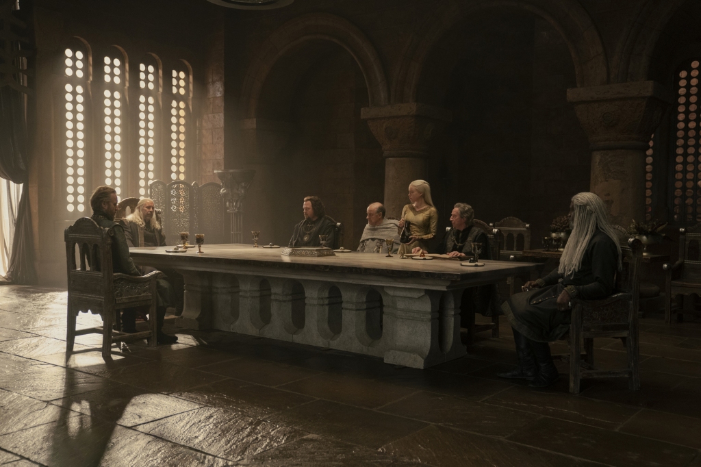 King Viserys (Paddy Considine), Princess Rhaenyra (Milly Alcock) and the rest of the Small Council sitting at a long table. 