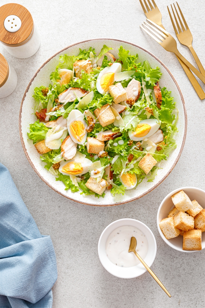 A caesar salad with grilled chicken breast, hard-boiled egg, croutons, parmesan cheese, green lettuce and dressing.
