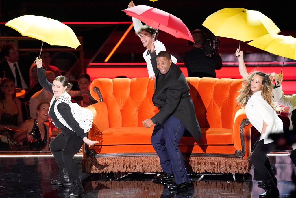 Kenan Thompson sits on an orance couch with an open umbrella onstage. 