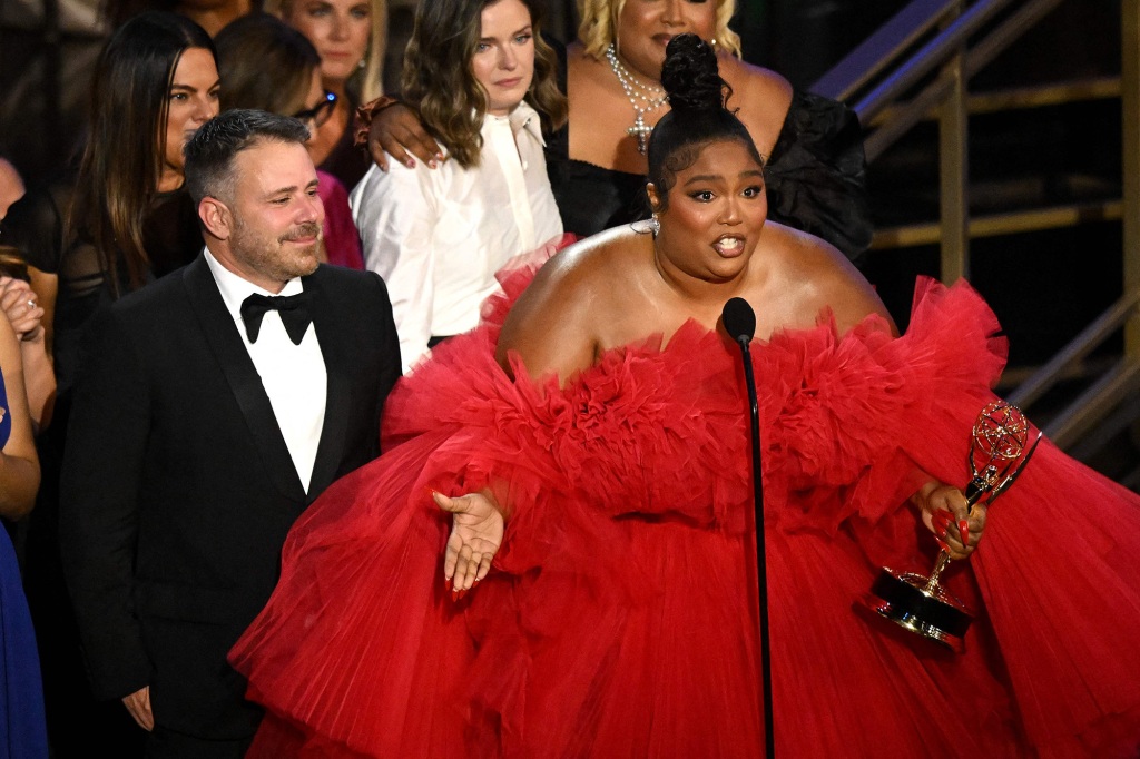 Lizzo accepting her Emmy award. She's wearing a bright red dress and talking into the microphone and holding her Emmy in her left hand.
