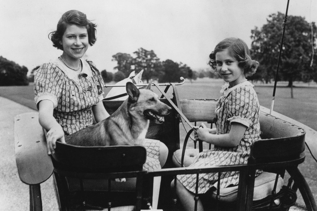 Black and white photo showing a younger Queen Elizabeth and her younger sister Princess Margaret sitting in carriage with a corgi in 1940.