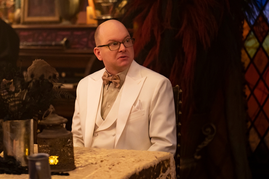 Mark Proksch as Colin Robinson in his boring energy vampire form from Season 3. He's sitting at a table an wearing a white suit with a white vest and a brown shirt and bowtie. He's looking off camera and is wearing glasses.