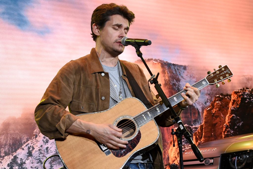 John Mayer sings into a microphone while holding a guitar.