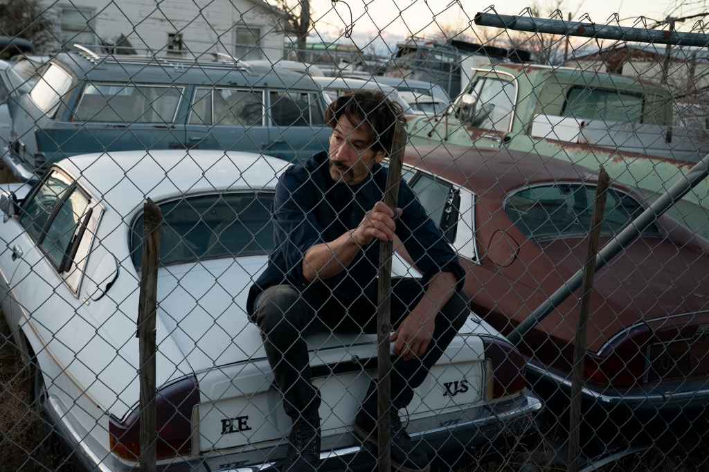 Jon Bernthal sitting on the hood of a  car behind a chain link fence. 