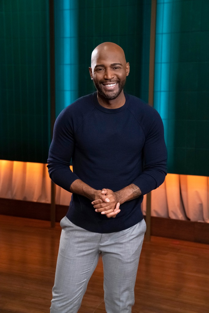 Photo of Karamo Brown on the set of his talk show. He's looking directly at the camera with a smile and is wearing a blue pullover sweater and grey pants. His hands are clasped in front of him.