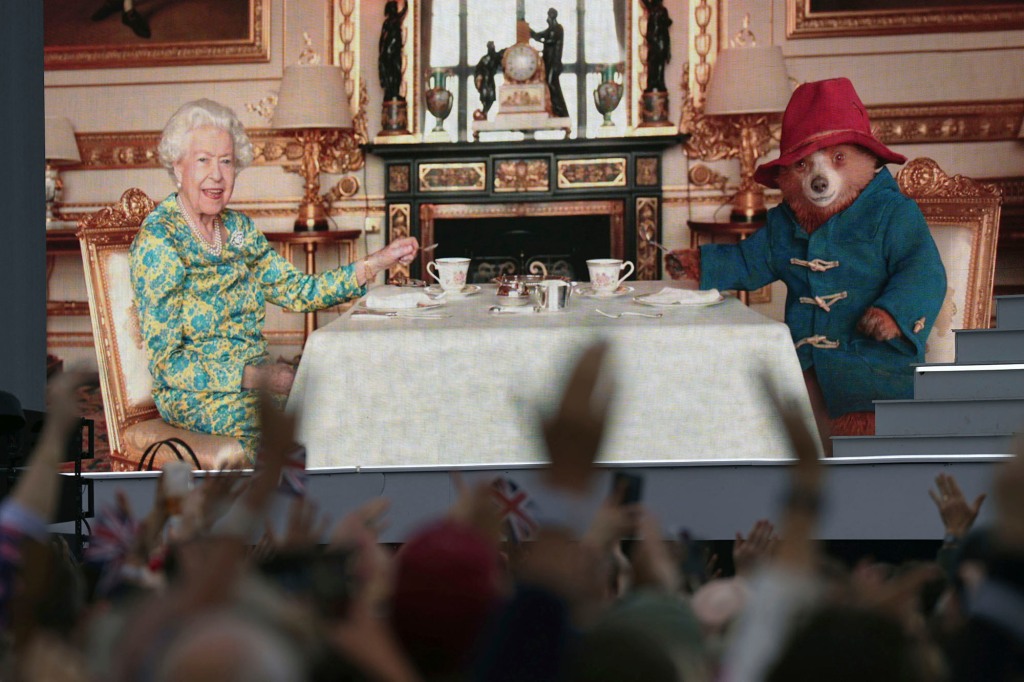 The crowd watch a film of Queen Elizabeth II having tea with Paddington Bear on a big screen at the Platinum Jubilee concert taking place in front of Buckingham Palace, London, Saturday June 4, 2022, on the third of four days of celebrations to mark the Platinum Jubilee. The events over a long holiday weekend in the U.K. are meant to celebrate Queen Elizabeth II’s 70 years of service. (Victoria Jones/PA via AP)