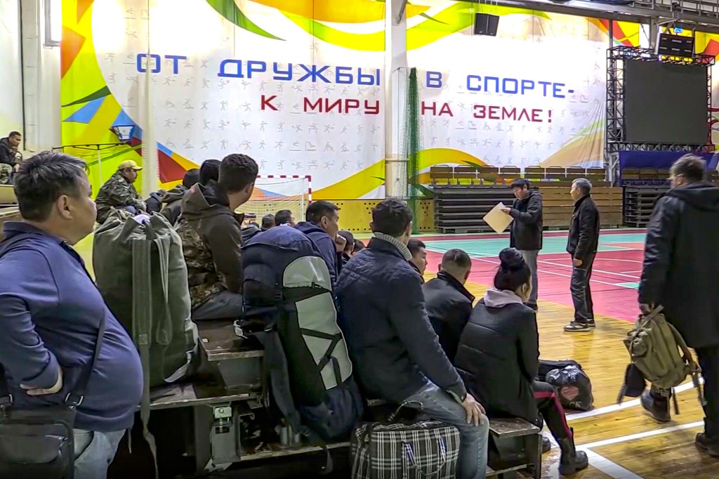 Russian draftees gather inside an indoor stadium turned into a collection center. The words in the background read "From friendship in sports to peace on earth!" 