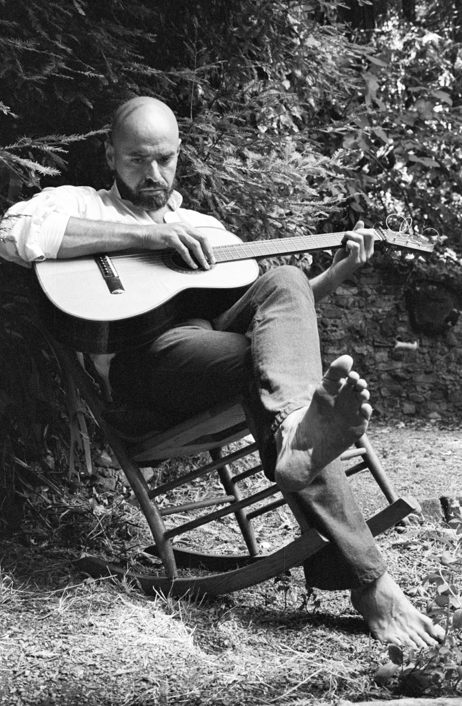 Shel Silverstein, barefoot, holds a guitar in a rocking chair. 