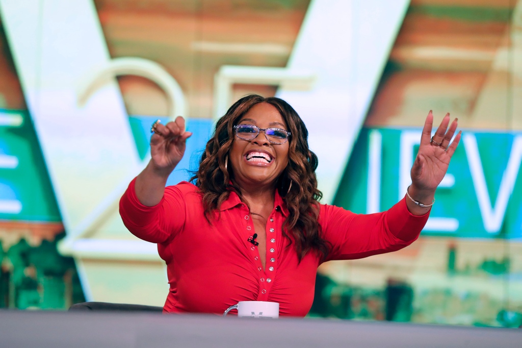 Sherri Shepherd sitting at a desk smiling with her arms up. 