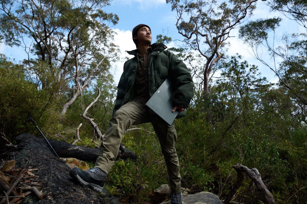 A user holds the Acer Swift Edge under their left arm and stands on a hill in an outdoor forest setting, looking over their left shoulder.