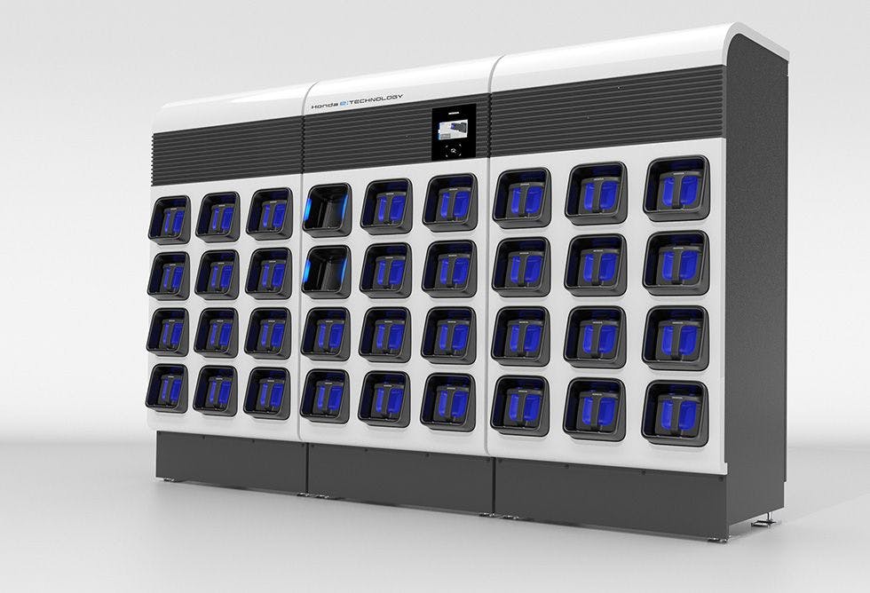 Three vending machine-like modules make up this expanded version of the Honda power pack exchanger, it has three square pack slots across and four down per unit, making it support 12 per unit and making this big version support 36. The machine is white and gray colored with a touch screen on the main central unit, and all the packs are blue.