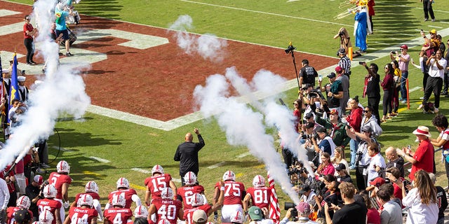 Head Coach David Shaw, wearing black, of the Stanford Cardinal leads his team onto the field before an NCAA Pac-12 college football game against the Arizona State Sun Devils on Oct. 22, 2022 at Stanford Stadium in Palo Alto, California; visible players include Brycen Tremayne carrying the American flag.  