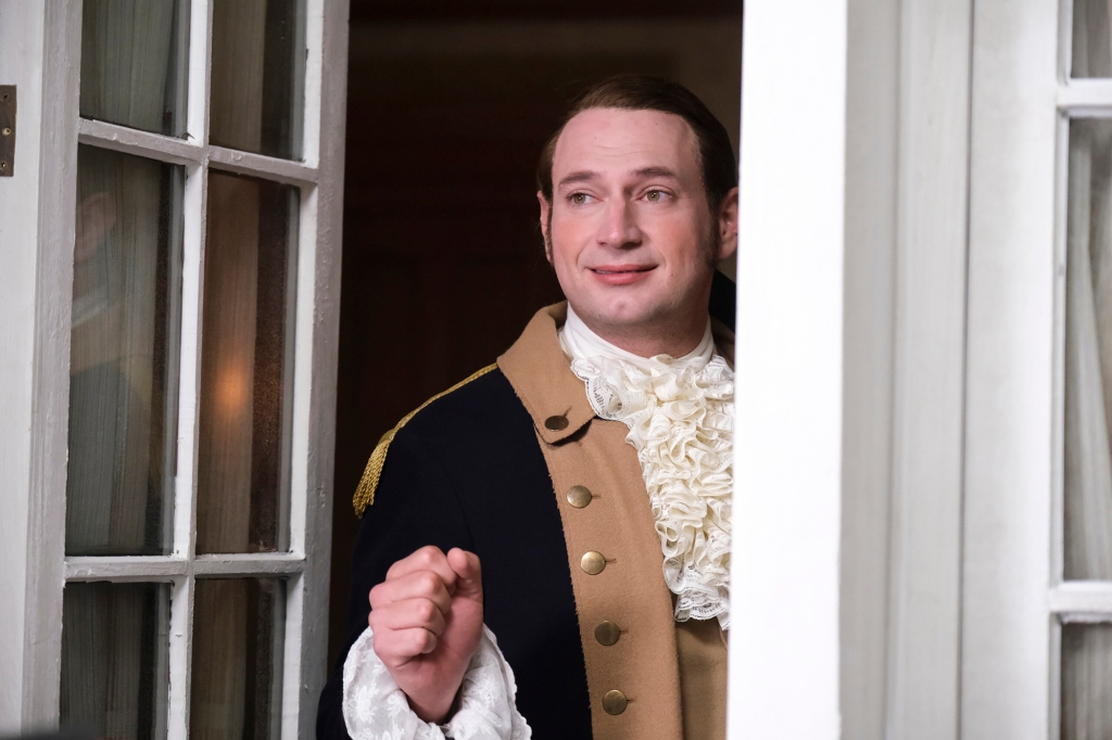 Brandon Scott Jones as Isaac. He's wearing a Revolutionary War uniform and is looking out the window and smiling.