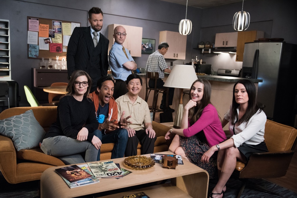 Gillian Jacobs, Joel McHale, Danny Pudi, Jim Rash, Ken Jeong, Alison Brie, Paget Brewster in "Community" stand in a living room, some sitting on a couch.