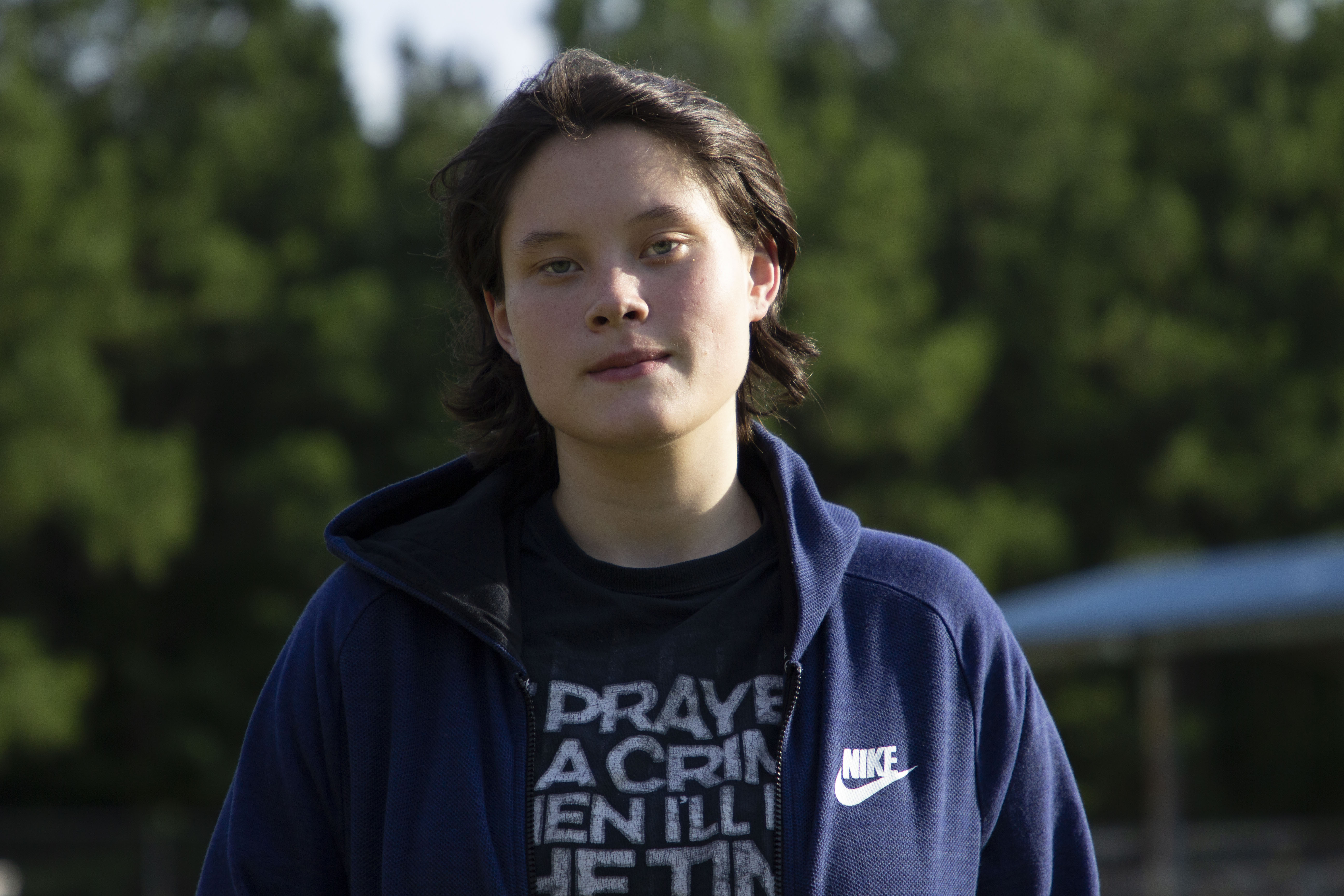 Cora Sepulvado, standing with a backdrop of trees behind her, looking at the camera and wearing a dark T-shirt and blue sweatshirt.