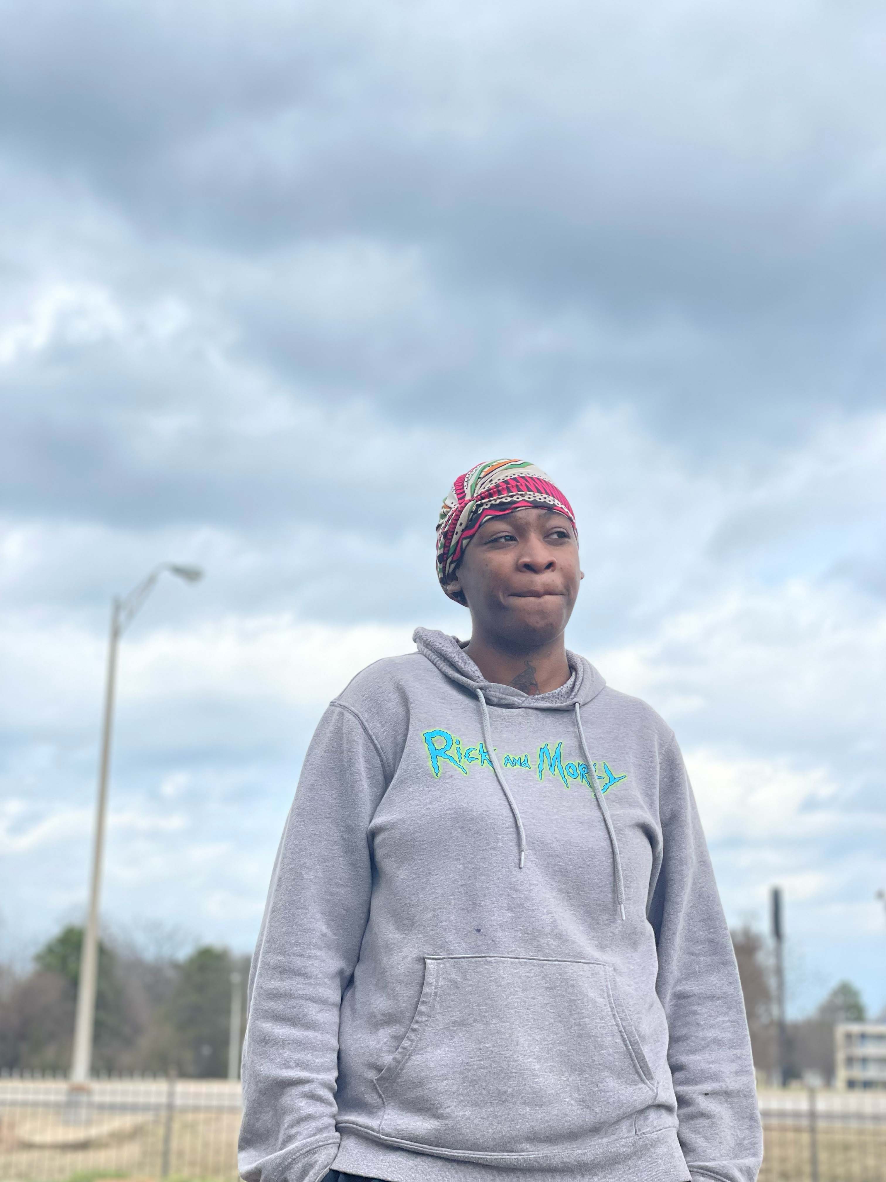 Doreisha Martin standing against a blue sky with clouds, looking to the right. She wears a gray hooded sweatshirt and a colorful headwrap.