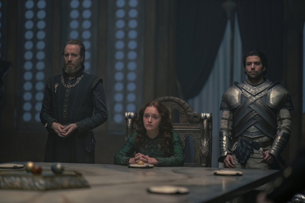 Otto Hightower (Rhys Ifans) Alicent (Olivia Cooke) and Criston Cole (Fabien Frankel) in "House of the Dragon" sit at a table looking grim. 