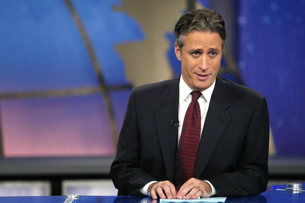 Photo of a younger Jon Stewart sitting behind "The Daily Show" desk. He's wearing a dark suit and red tie and is hair is much darker than it is now.