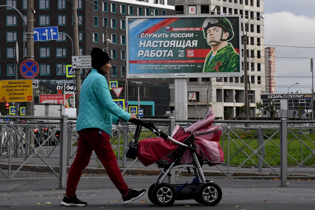 a woman pushes a stroller past a sign reading "Serving Russia is a real job"