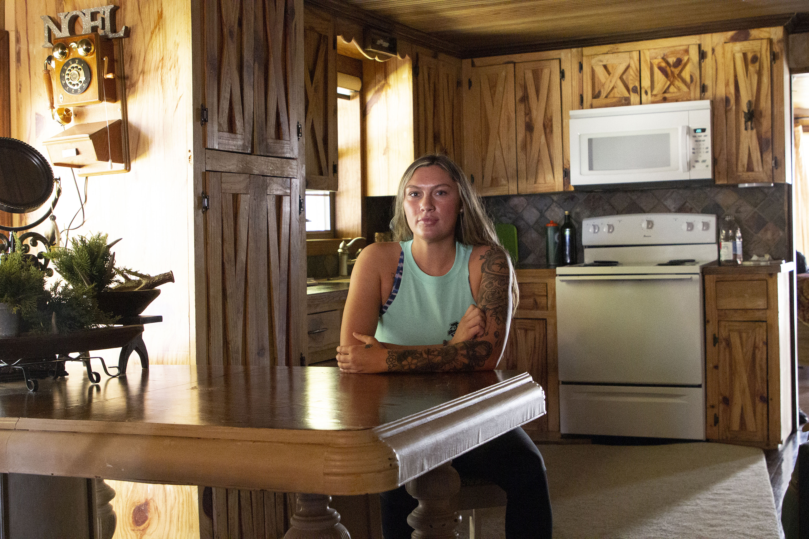 Natalee Brannon sitting at a kitchen table, looking at the camera. She wears an aqua tank top and has tattoos on her left arm.