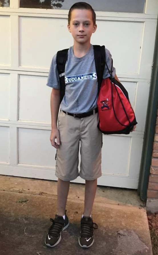 Solan Peterson standing in front of a white garage door, looking at the camera with a neutral expression. He wears a gray Buccaneers T-shirt, khaki shorts and a backpack and holds a red bag.